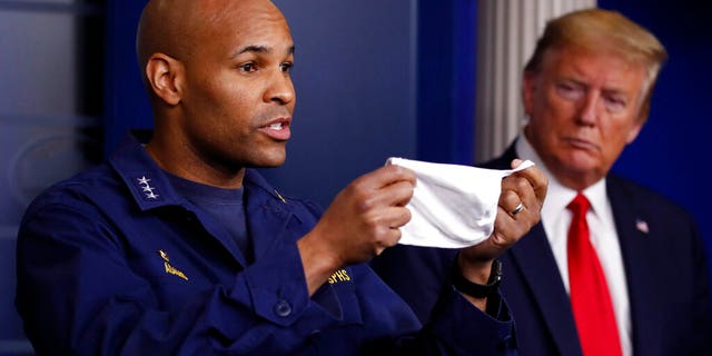 In this April 22, photo President Trump watches as U.S. Surgeon General Jerome Adams holds up his face mask at a coronavirus briefing in the White House. Adams was cited for being in a closed Hawaii park in August while in the islands helping with surge testing amid a spike in coronavirus cases. (AP Photo/Alex Brandon, File)