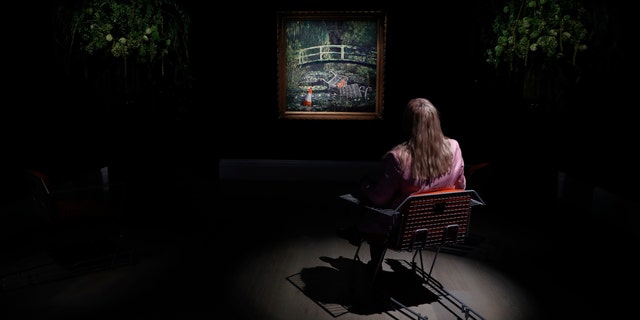 A member of staff poses in front of an artwork by Banksy entitled 'Show Me The Monet" at Sotheby's auction house in London, Friday, Oct. 16, 2020.