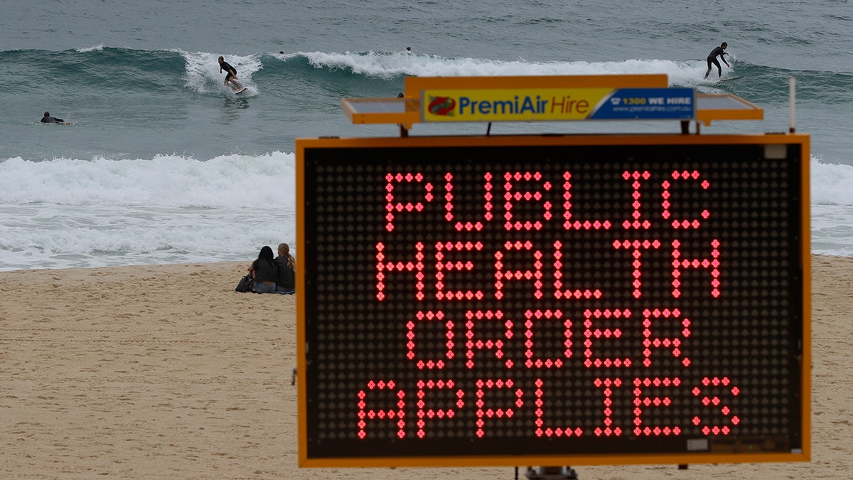 Surfers ride a wave past a sign at a beach in Sydney, Australia, Saturday, Dec. 19, 2020. Sydney's northern beaches will enter a lockdown similar to the one imposed during the start of the COVID-19 pandemic in March as a cluster of cases in the area increased to more than 40. (AP Photo/Mark Baker)