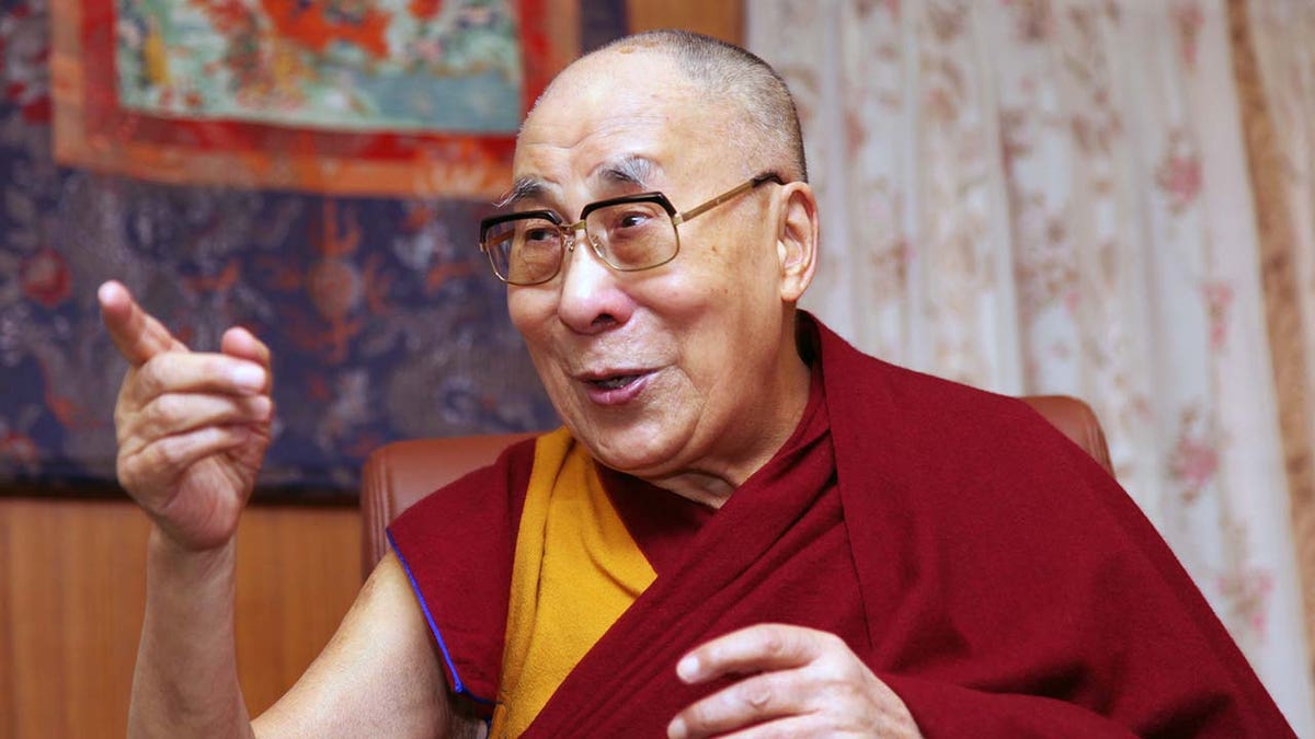 Dalai Lama points off camera wearing eyeglasses and his traditional wear