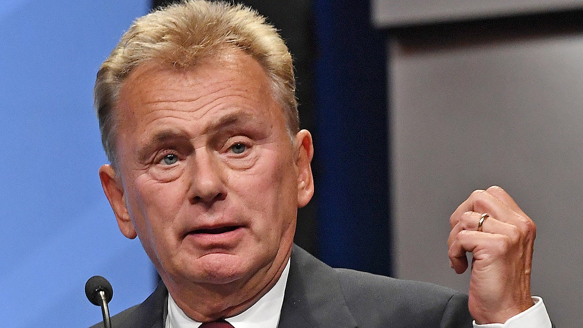 Pat Sajak in a grey suit looks perplexed with his hand partially raised