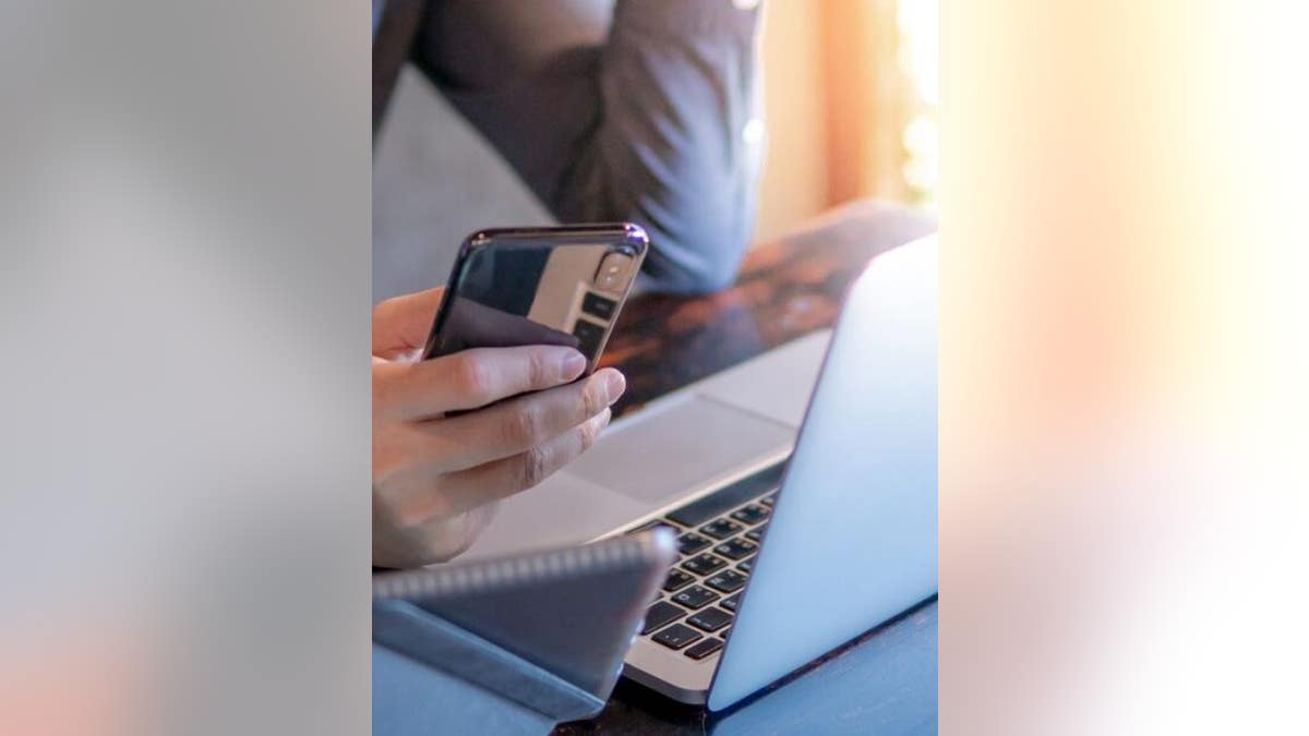 A man uses his Iphone and laptop computer at his desk.