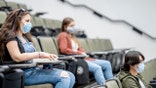 Multi-ethnic group of students wearing protective face masks while sitting in a lecture hall sitting 2 meters apart.