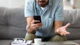 Close up confused man having problem with broken not working phone, annoyed frustrated male student receiving bad news, reading unpleasant email, sitting on sofa, looking at screen, spam message