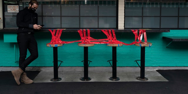 A man looks at his phone next to stools taped off to prevent diners from sitting on them in Los Angeles on Monday, Dec. 7, 2020. (AP Photo/ Jae C.Hong, File)