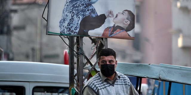 A man wearing a face mask to protect against the coronavirus stands next to a poster promoting a polio campaign at a market in Peshawar, Pakistan, Dec. 14, 2020. The task of vaccinating millions of people in poor and developing countries against COVID-19 faces monumental obstacles, and it's not just a problem of affording and obtaining doses. In parts of Pakistan, it can be outright deadly: More than 100 workers in polio vaccination campaigns have been killed since 2012, as militants spread fears that the inoculations are an anti-Islam plot. (AP Photo/Muhammad Sajjad)