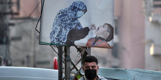 A man wearing a face mask to protect against the coronavirus stands next to a poster promoting a polio campaign at a market in Peshawar, Pakistan, Dec. 14, 2020. The task of vaccinating millions of people in poor and developing countries against COVID-19 faces monumental obstacles, and it's not just a problem of affording and obtaining doses. In parts of Pakistan, it can be outright deadly: More than 100 workers in polio vaccination campaigns have been killed since 2012, as militants spread fears that the inoculations are an anti-Islam plot. (AP Photo/Muhammad Sajjad)