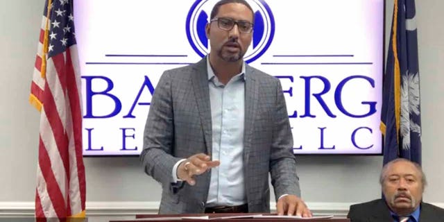 In this screengrab from video provided by Bamberg Law, LLC, attorney Justin Bamberg, standing, speaks at a news conference as plaintiff Jethro DeVane, seated at right, listens, Tuesday, Dec. 22, 2020, in Orangeburg, S.C. (Courtesy of Bamberg Law, LLC via AP)