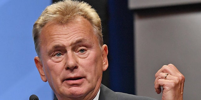 Pat Sajak in a grey suit looks perplexed with his hand partially raised