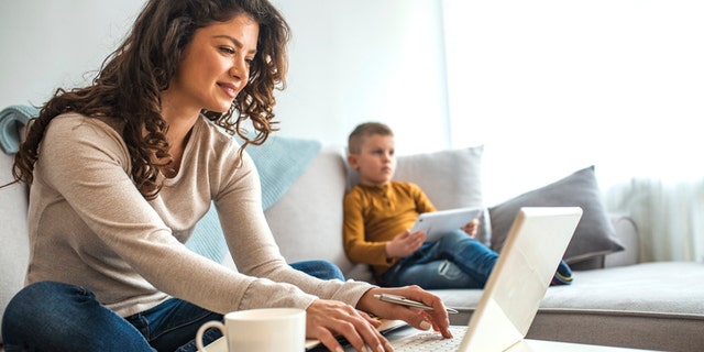 A mother works from home as her son holds a tablet on the sofa. (iStock)