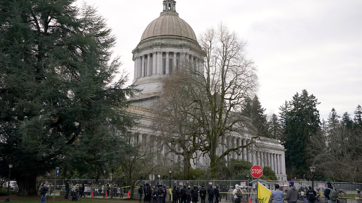 A group of anti-fascist protesters dressed in black make a brief stop outside a perimeter fence being watched by National Guard members and Washington State Patrol troopers, Sunday, Jan. 10, 2021, at the Capitol in Olympia, Wash. Washington Gov. Jay Inslee activated members of the National Guard this week to work with the Washington State Patrol to protect the Capitol campus. The group, part of a larger protest that was taking place in downtown Olympia, left after several minutes of exchanging words with troopers and guard members. (AP Photo/Ted S. Warren)