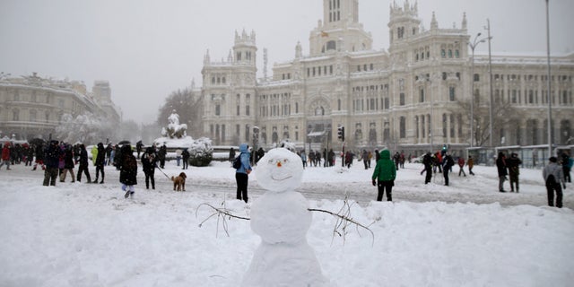 People walk past the Cibeles monument, left, in front of the City Hall during a heavy snowfall in downtown Madrid, Spain, Saturday, Jan. 9, 2021. A persistent blizzard has blanketed large parts of Spain with 50-year record levels of snow, halting traffic and leaving thousands trapped in cars or in train stations and airports that suspended all services as the snow kept falling on Saturday. (AP Photo/Andrea Comas)