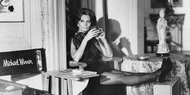 Actress Sophia Loren, with a drink in hand and a portable fan offering her relief from the heat, cools her heels during a break in the filming of the movie 'Firepower,' directed by Michael Winner.