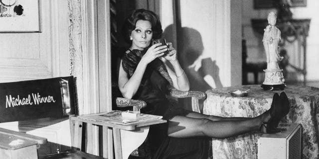 Actress Sophia Loren, with a drink in hand and a portable fan offering her relief from the heat, cools her heels during a break in the filming of the movie 'Firepower,' directed by Michael Winner.