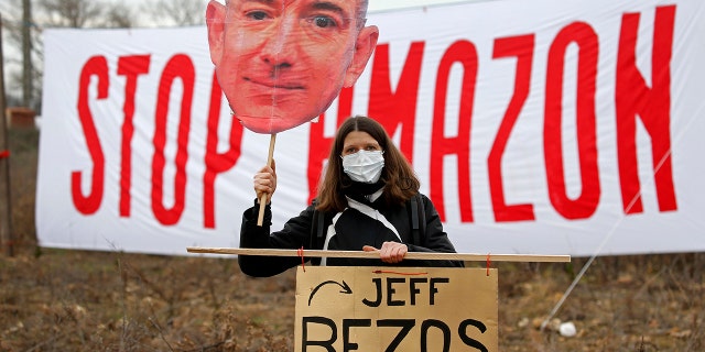 OURNES, FRANCE - 일월 30: An opponent holds a cutout sign depicting Amazon CEO Jeff Bezos, in front of a banner reading "Stop Amazon" during a rally against plans to build a giant Amazon warehouse on January 30, 2021 in Fournes (Gard), 프랑스. The project to set up a huge Amazon warehouse by the e-commerce giant in Fournes, near the Pont du Gard, faces strong opposition. The demonstrators oppose the "betonization" 의 14 hectares of land a few kilometers from the Pont du Gard, (classified as a historical monument as a UNESCO World Heritage Site). (Photo by Chesnot/Getty Images)