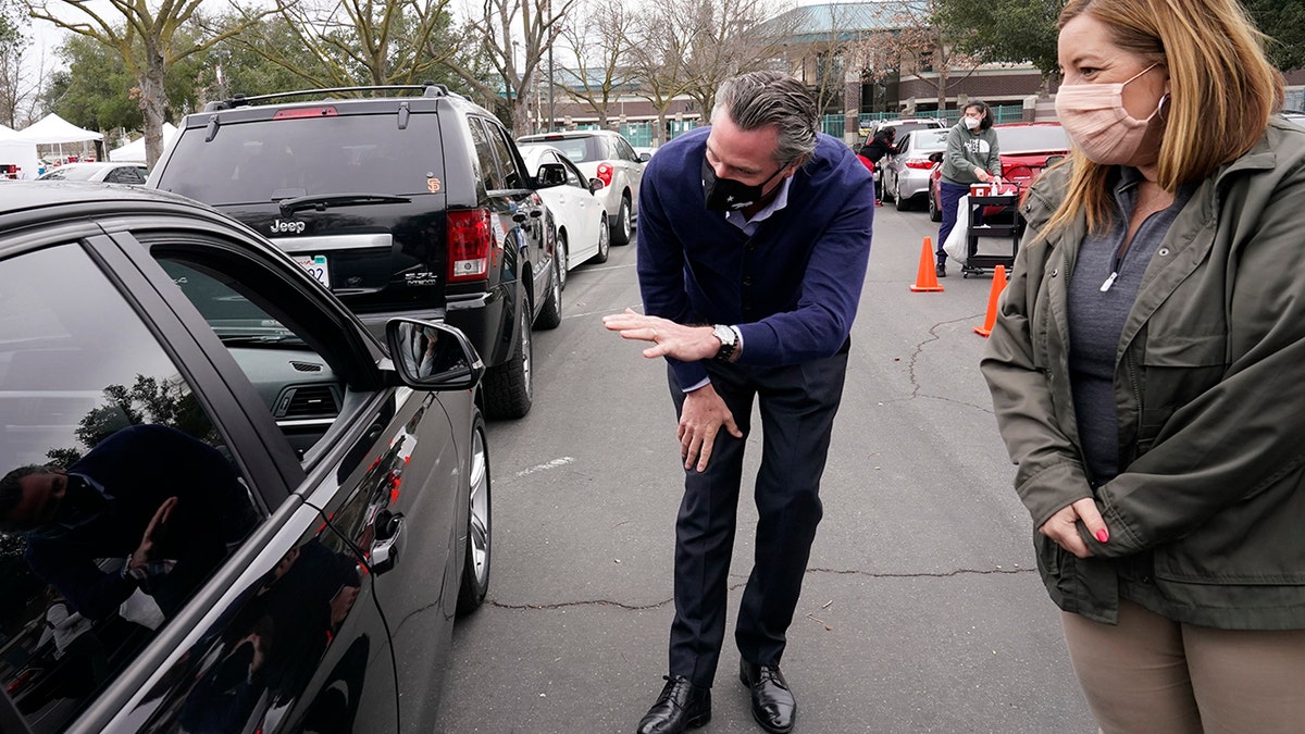 Gov. Gavin Newsom leans over to talk with a person waiting to get a COVID-19 vaccination at a drive-thru vaccination center at Natomas High School in Sacramento, Calif., Thursday, Feb. 11, 2021. Appointments were needed for the 1,000 vaccinations to be administered for those 65 and over, first responders, health workers, teachers, food and agricultural employers. Called an equitable distribution site, it prioritized those disproportionally impacted by COVID-19, was collaboration between the Natomas' Unified School District, Sacramento County Public Health Department and Sacramento Vice Mayor Angelique Ashby, behind Newsom, who represents the area. (AP Photo/Rich Pedroncelli)