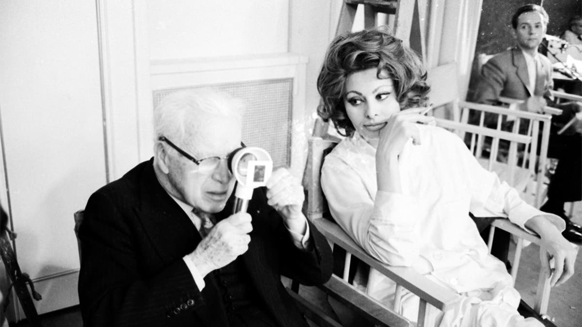 Charlie Chaplin looking a film negative through a magnifying glass while sitting close to actress Sophia Loren during filming on the set of the movie 'A Countess from Hong Kong' at the Pinewood Studios in London, 1966