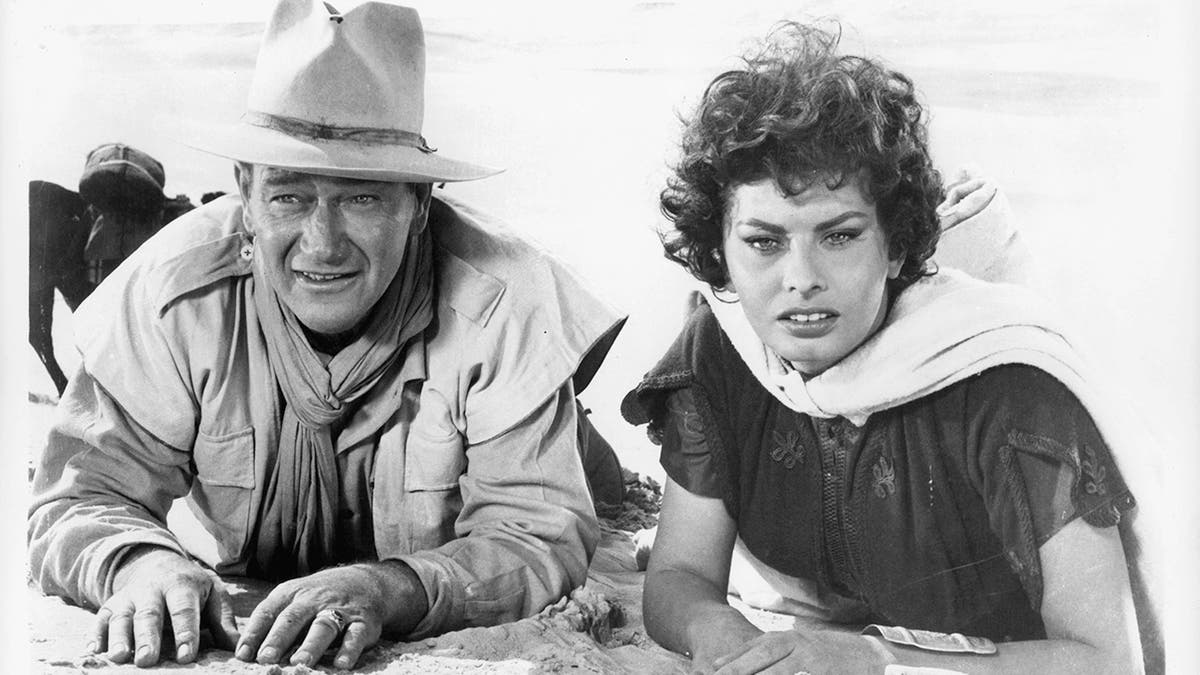 John Wayne and Sophia Loren laying in the sand in a scene from the film 'Legend Of The Lost', 1957. 