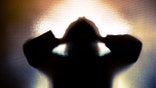 Colour backlit image of the silhouette of a woman with her hands on her head in a gesture of despair. The silhouette is distorted, and the arms elongated, giving an alien-like quality. The image is sinister and foreboding, with an element of horror. It is as if the woman is trying to escape from behind the glass. Horizontal image with copy space.