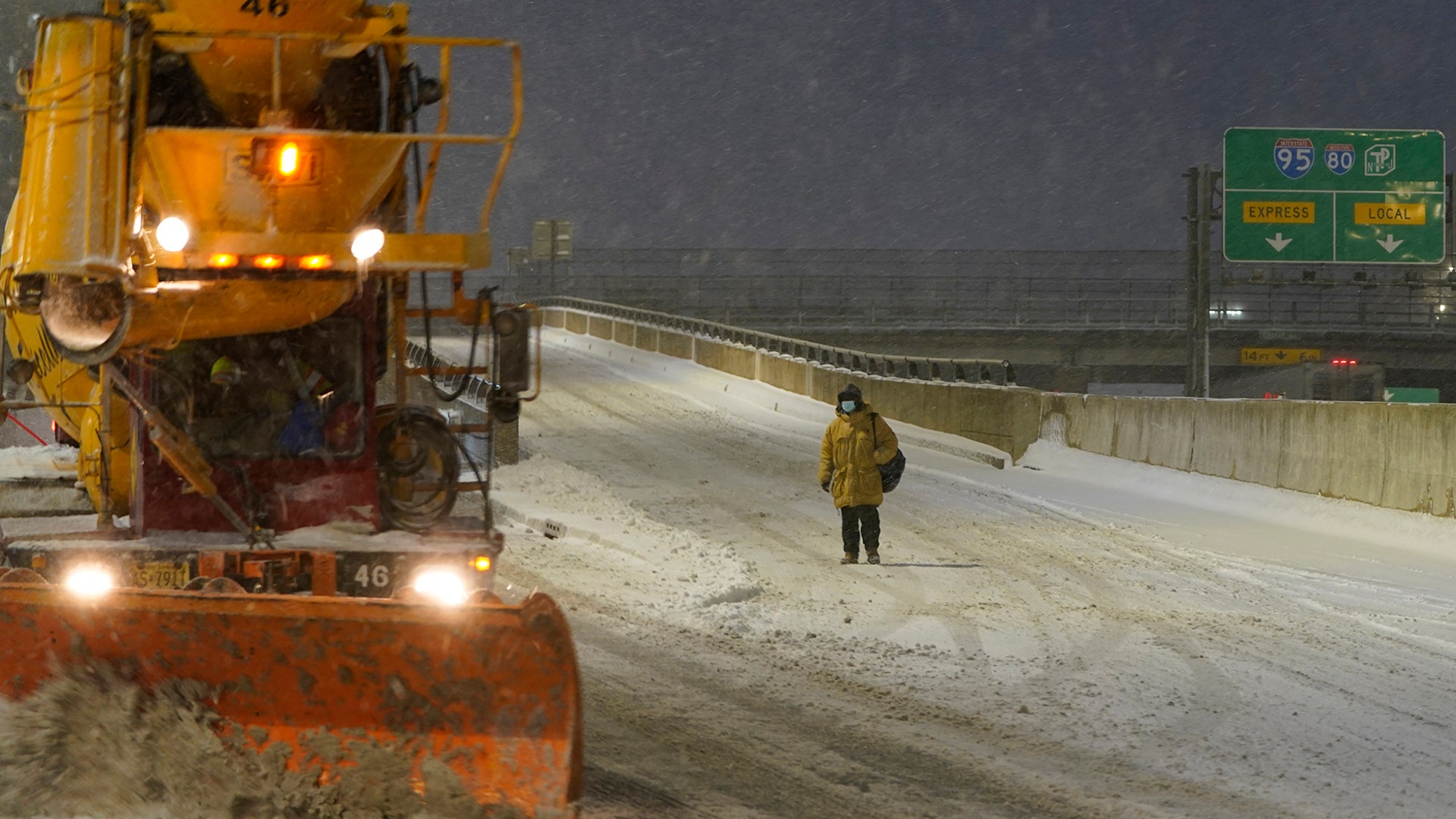 A person waits for a snow plow to pass before crossing the street in Fort Lee, N.J., Monday, Feb. 1, 2021. Snowfall is picking up in the Northeast as the region braced for a whopper of a storm that could dump well over a foot of snow in many areas, create blizzard-like conditions and cause travel problems for the next few days. (AP Photo/Seth Wenig)