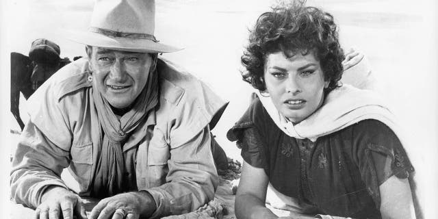 John Wayne and Sophia Loren laying in the sand in a scene from the film 'Legend Of The Lost', 1957.