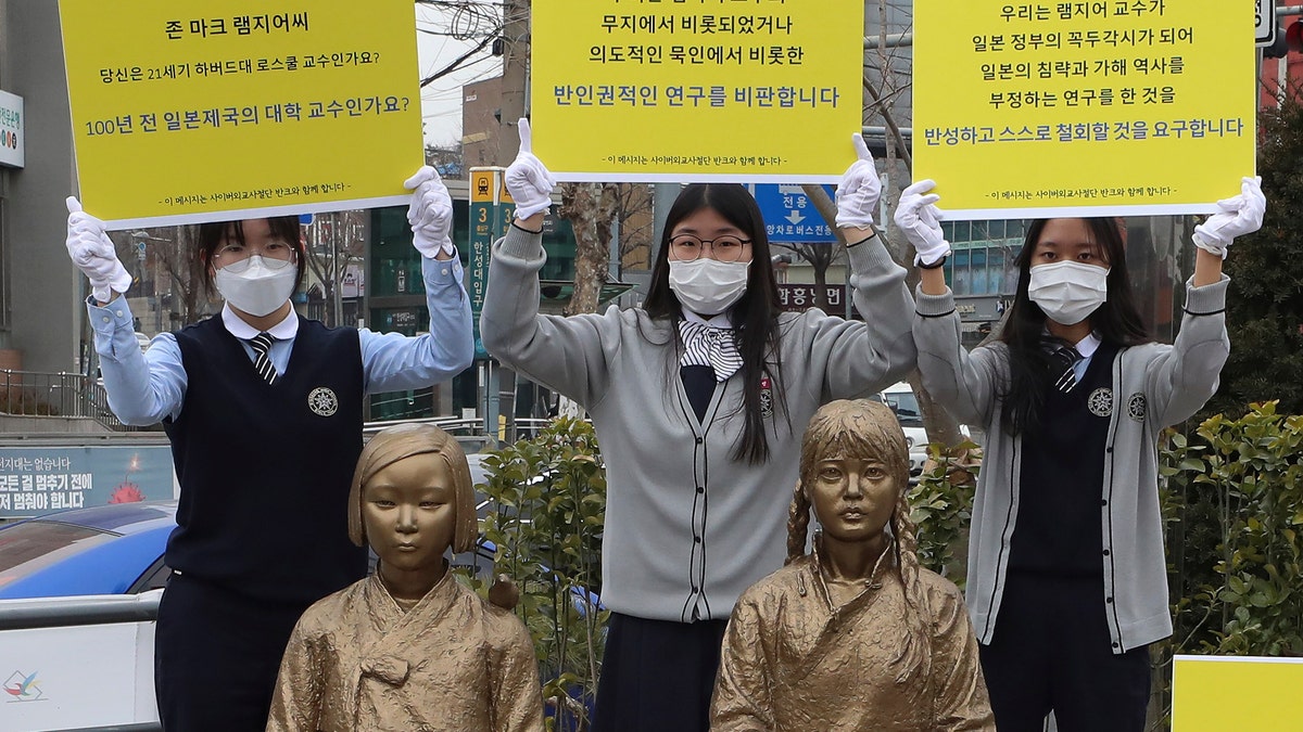 In this Feb. 25, 2021, photo, high school students hold up banners to protest a recent academic paper by Harvard University professor J. Mark Ramseyer, behind statues symbolizing wartime sex slaves in Seoul, South Korea. The signs read: "J. Mark Ramseyer, are you a 21st century professor at Harvard? Are you a university professor in the Japanese Empire 100 years ago? We criticize anti-human rights research." (Lee Jung-hoon/Yonhap via AP)