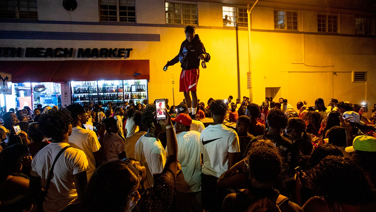 A man stands on a car as crowds defiantly gather in the street while a speaker blasts music an hour past curfew in Miami Beach, Fla., on Sunday, March 21, 2021. An 8 p.m. curfew has been extended in Miami Beach after law enforcement worked to contain unruly crowds of spring break tourists. (Daniel A. Varela/Miami Herald via AP)