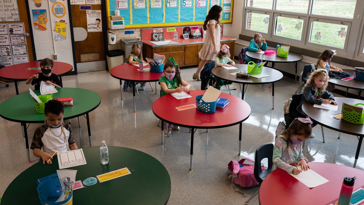 Students seen sitting a tables in a school classroom as a teacher walks through the class