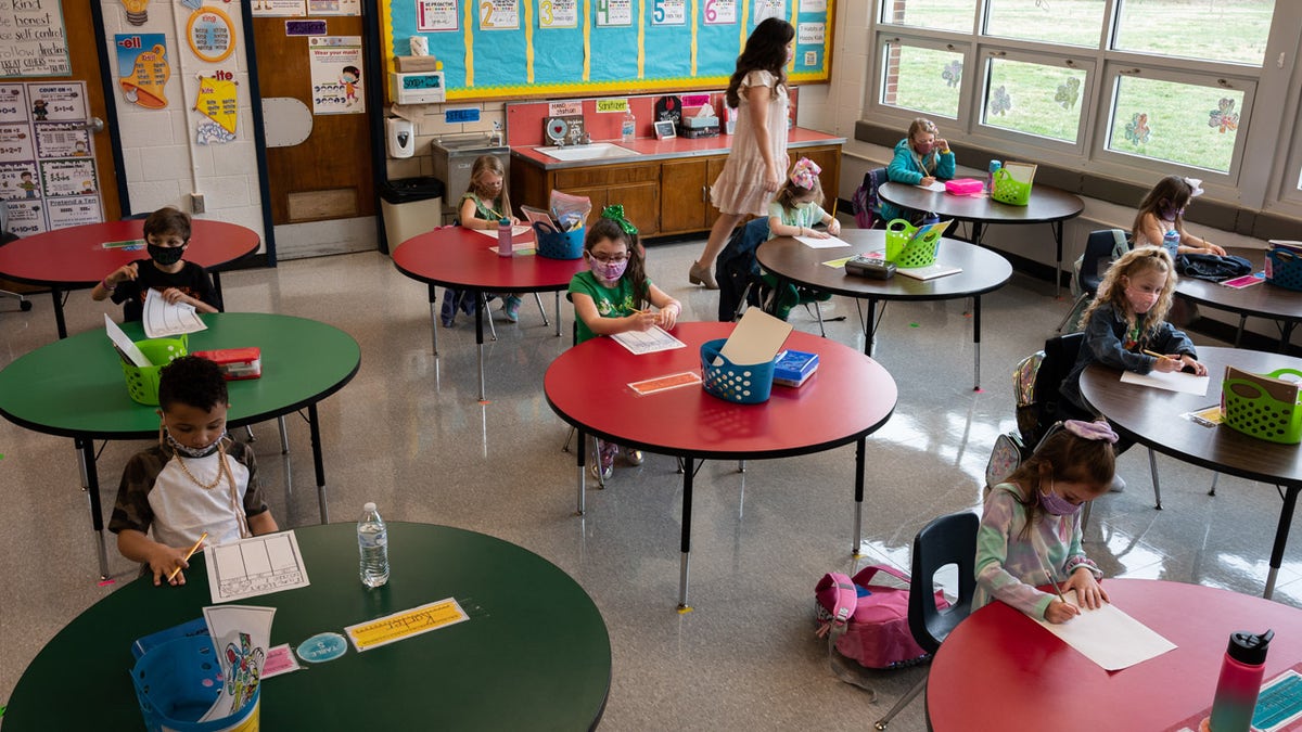 Students at tables in classroom, teacher walking in background