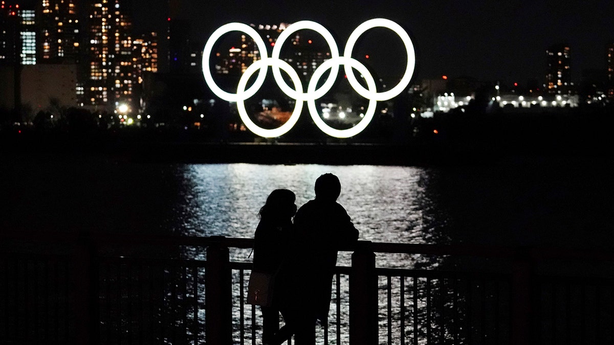 A man and a woman stand with the backdrop of the Olympic rings floating in the water in the Odaiba section in Tokyo, Wednesday, March 3, 2021. (AP Photo/Eugene Hoshiko)