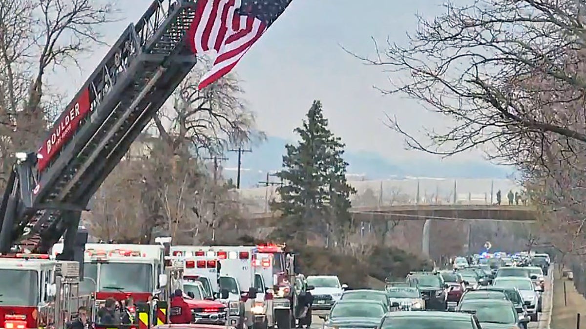 Vehicles pass under a giant American flag stationed alongside a road in Boulder, Colo., during a procession for a fallen police officer.