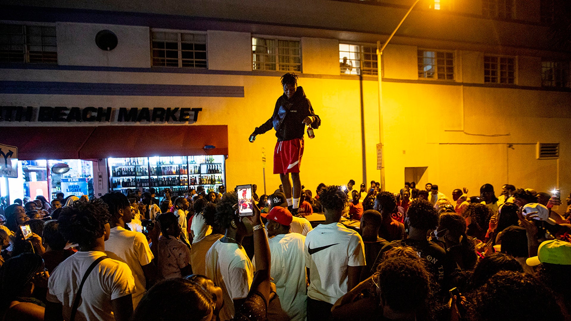 A man stands on a car as crowds defiantly gather in the street while a speaker blasts music an hour past curfew in Miami Beach, Florida, on Sunday, March 21, 2021. An 8 p.m. curfew has been extended in Miami Beach after law enforcement worked to contain unruly crowds of spring break tourists. (Daniel A. Varela/Miami Herald via AP)