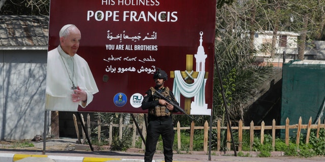 Security stand by a poster depicting Pope Francis, in Baghdad Iraq, Friday, March 5, 2021. Pope Francis heads to Iraq on Friday to urge the country's dwindling number of Christians to stay put and help rebuild the country after years of war and persecution, brushing aside the coronavirus pandemic and security concerns. (AP Photo/Andrew Medichini)