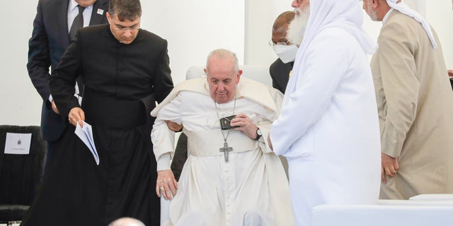 Pope Francis is helped standing during an interreligious meeting near the archaeological area of the Sumerian city-state of Ur, 20 kilometers south-west of Nasiriyah, Iraq, Saturday, March 6, 2021. Since the beginning of this trip to Iraq Francis has been limping from an apparent flare-up of sciatica nerve pain that makes walking difficult. Ur is considered the traditional birthplace of Abraham, the prophet common to Muslims, Christians and Jews.Francis traveled to the southern ruins of Ur on Saturday to reinforce his message of interreligious tolerance and fraternity during the first-ever papal visit to Iraq, a country riven by religious and ethnic divisions. (AP Photo/Andrew Medichini)