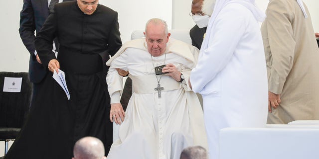 Pope Francis is helped standing during an interreligious meeting near the archaeological area of the Sumerian city-state of Ur, 20 kilometers south-west of Nasiriyah, Iraq, Saturday, March 6, 2021. Since the beginning of this trip to Iraq Francis has been limping from an apparent flare-up of sciatica nerve pain that makes walking difficult. Ur is considered the traditional birthplace of Abraham, the prophet common to Muslims, Christians and Jews.Francis traveled to the southern ruins of Ur on Saturday to reinforce his message of interreligious tolerance and fraternity during the first-ever papal visit to Iraq, a country riven by religious and ethnic divisions. (AP Photo/Andrew Medichini)