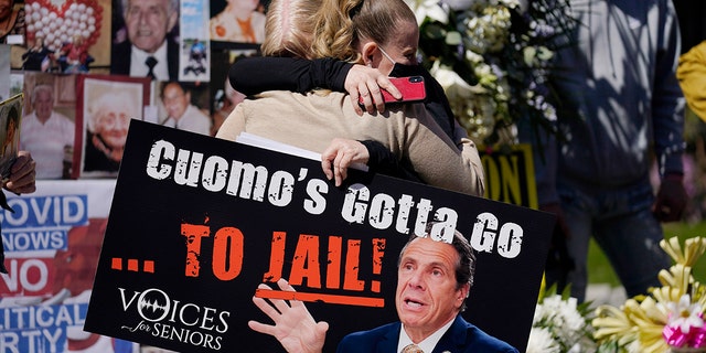 A woman holding a sign hugs another woman in front of a section of the memorial park at a conference in New York, Sunday, March 21, 2021. Families gathered to mourn but they wanted to a lawsuit and claim from Gov. Andrew Cuomo. (AP Photo/Seth Wenig)