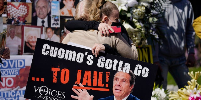 A woman holding a sign hugs another woman in front of a section of a memorial wall at a news conference in New York, Sunday, March 21, 2021. The families  gathered to grieve but want an investigation into and accountability from Gov. Andrew Cuomo. (AP Photo/Seth Wenig)