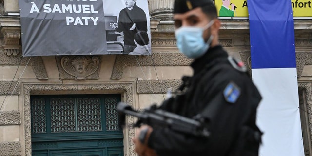 A French police officer stands next to a portrait of French teacher Samuel Paty on display on the facade of the Opera Comedie in Montpellier on October 21, 2020, during a national homage to the teacher who was beheaded for showing cartoons of the Prophet Mohamed in his civics class. (Photo by Pascal GUYOT / AFP) (Photo by PASCAL GUYOT/AFP via Getty Images)