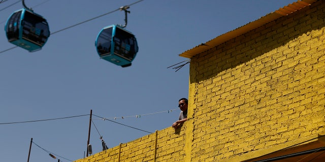 A man stands on a rooftop as cable cars overhead run between the Campos Revolucion and Tlalpexco stations. 