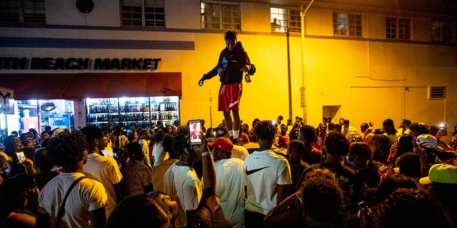 A man stands on a car as crowds defiantly gather in the street while a speaker blasts music an hour past curfew in Miami Beach, Fla., on Sunday, March 21, 2021. An 8 p.m. curfew has been extended in Miami Beach after law enforcement worked to contain unruly crowds of spring break tourists. (Daniel A. Varela/Miami Herald via AP)