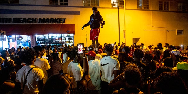 A man stands on a car as crowds defiantly gather in the street while a speaker blasts music an hour past curfew in Miami Beach, Fla., on Sunday, March 21, 2021. An 8 p.m. curfew has been extended in Miami Beach after law enforcement worked to contain unruly crowds of spring break tourists. (Daniel A. Varela/Miami Herald via AP)
