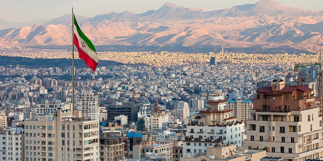 Waving Iran flag above skyline of Tehran at sunset. 