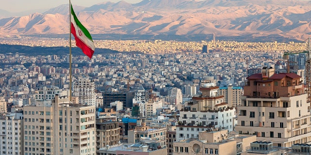 Waving Iran flag above skyline of Tehran at sunset.