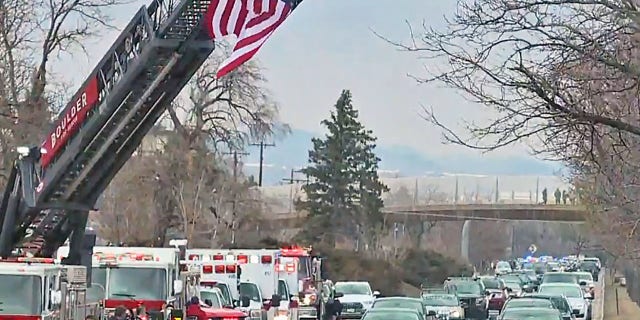Vehicles pass under a giant American flag stationed alongside a road in Boulder, Colo., during a procession for a fallen police officer.