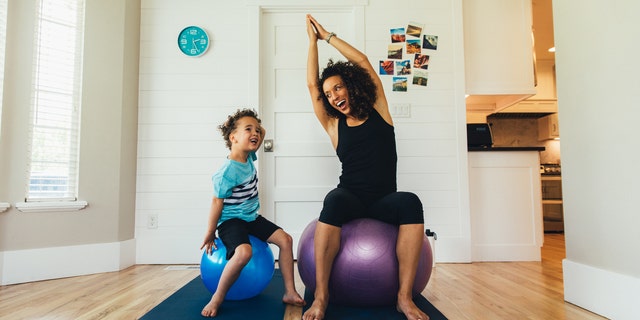 A mother exercises on a fitness ball with her young son inside their home. She is teaching the boy the importance of a healthy lifestyle by proper stretching and exercising.