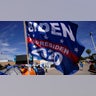 A woman passes a donated campaign flag for President Joe Biden at a makeshift camp of migrants at the border port of entry leading to the United States, Wednesday, March 17, 2021, in Tijuana, Mexico. The migrant camp shows how confusion has undercut the message from President Joe Biden that it’s not the time to come to the United States. Badly misinformed, some 1,500 migrants set up tents across the border from San Diego harbor in the hope that Biden will open entry briefly and without notice. (AP Photo/Gregory Bull)
