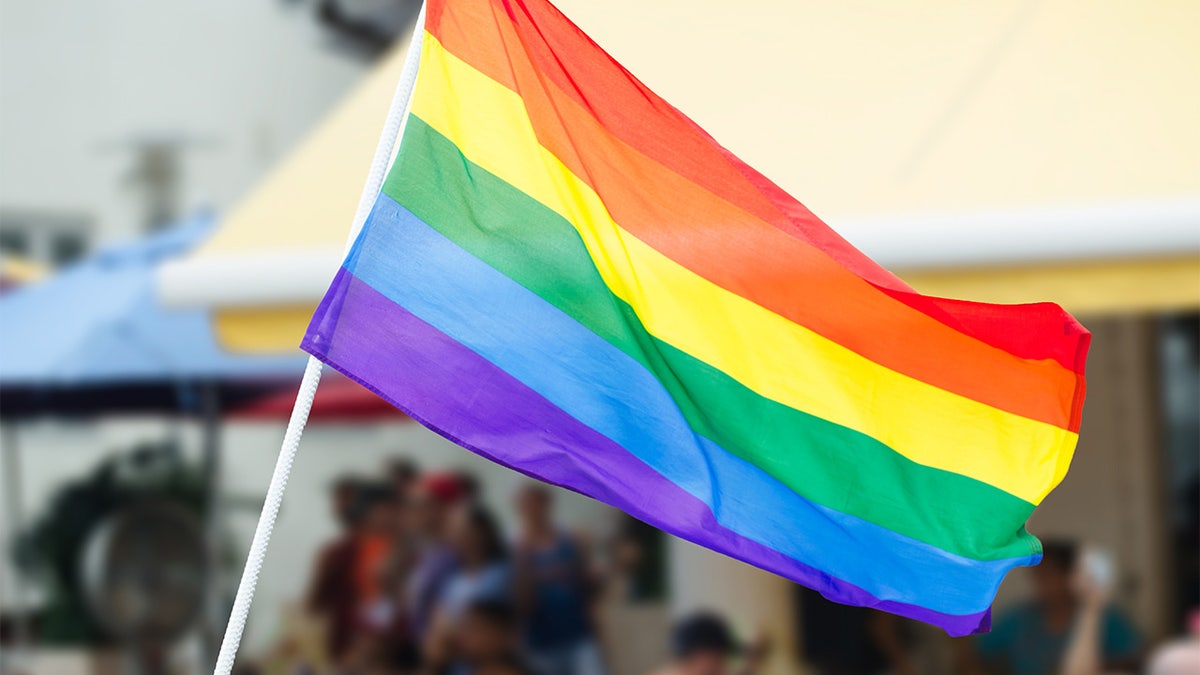 Rainbow flag waving on the street during a gay pride celebration with unrecognizable people lining the sidewalk in the background. U.S. embassies can now display the flag on the same pole as the American flag in June, during Pride month.