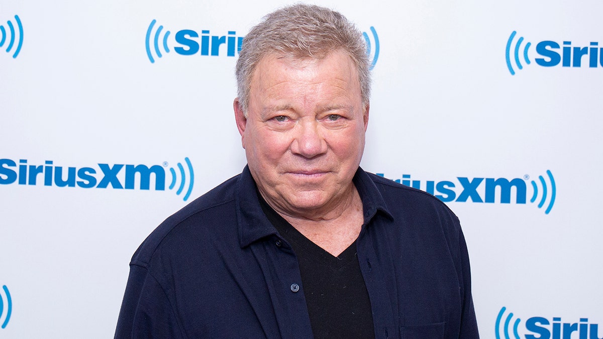 William Shatner wears a blue shirt open revealing a black undershirt in front of a SIRIUSXM backdrop