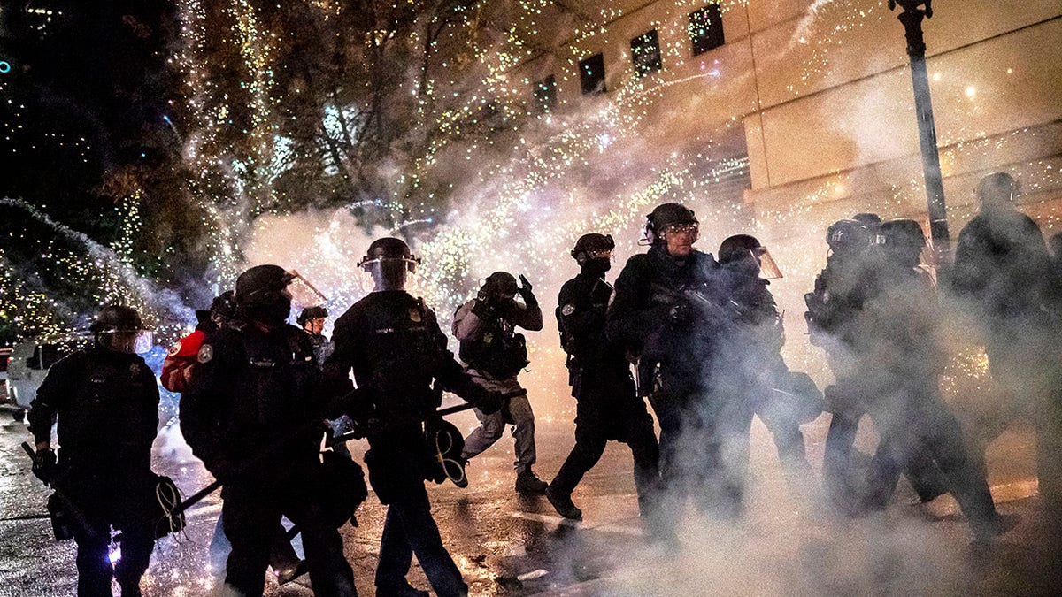 Police in riot gear walk through smoke with fireworks blasting behind them