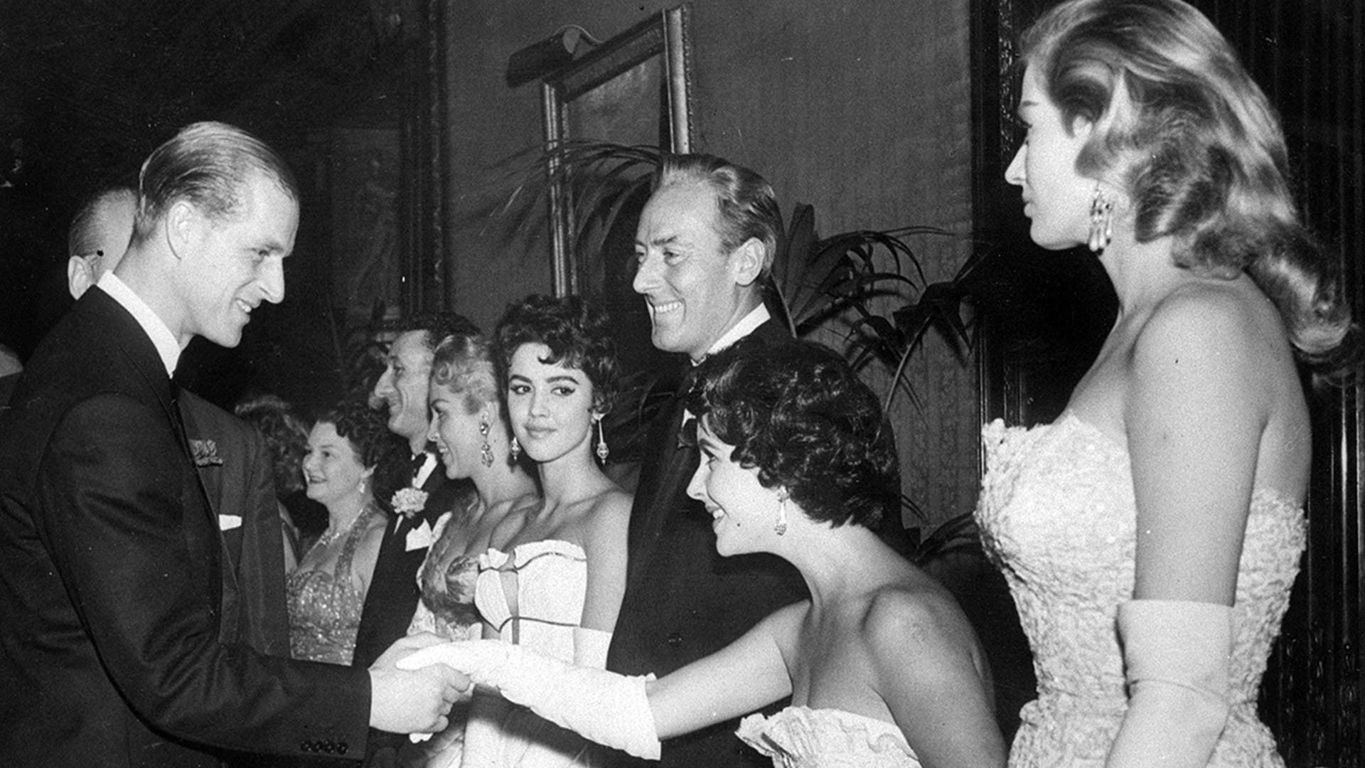FILE - In this Nov. 23, 1955 file photo, Elizabeth Taylor curtsies as she is greeted by the Duke of Edinburgh at the premiere of "Cockleshell Heroes" at the Empire Theater in London. To the left of Elizabeth Taylor is her husband Michael Wilding and at his right is actress Jackie Lane. Buckingham Palace says Prince Philip, husband of Queen Elizabeth II, has died aged 99. (AP Photo/File)